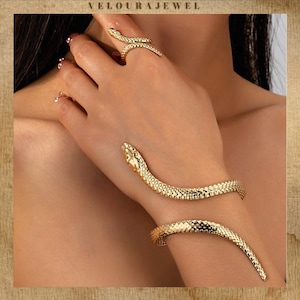 May include: Gold-coloured snake jewellery set, comprising a ring and a bracelet. The ring encircles a finger, and the bracelet wraps around the wrist. The jewellery features a textured, scale-like pattern. The brand name "VELOURAJEWEL" is at the top.