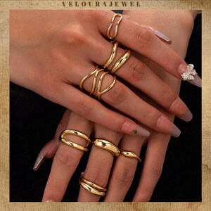 May include: A selection of gold rings in diverse styles, including a double band, curved bands, and abstract shapes. The rings are worn on multiple fingers, highlighting their designs. The brand name "VELOURAJEWEL" is visible at the top of the image.