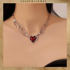 Puede incluir: Collar plateado con un colgante en forma de corazón con centro rojo. El diseño incluye acentos de manos esqueléticas a ambos lados del corazón, con la marca "VELOURAJEWEL" en la parte superior.