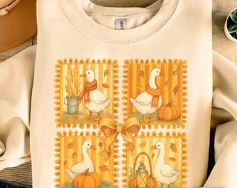 May include: Cream-colored sweatshirt featuring a fall-themed design. The design includes four framed illustrations of white geese wearing orange scarves, pumpkins, and autumn leaves. A large orange bow is centered below the illustrations.