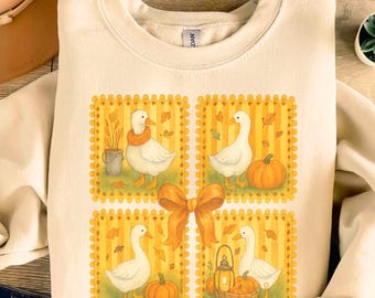 May include: Cream-colored sweatshirt featuring a whimsical fall design. The design includes four square panels with illustrations of white geese, pumpkins, and autumn leaves. A large orange bow is centered below the panels, adding a decorative touch.