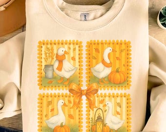 May include: Cream-colored sweatshirt featuring four square illustrations of geese in autumn settings. Each goose wears an orange scarf and is surrounded by pumpkins, leaves, and a lantern. A large orange bow is centered below the squares.