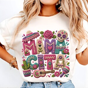 May include: Cream-colored t-shirt featuring the words "MAMA CITA" in colorful, textured letters. The design includes a sombrero, flowers, a sugar skull, a cactus, a guitar, and a maraca, all in a vibrant, crocheted style.