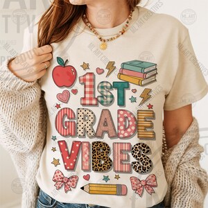 May include: Cream-colored t-shirt with the text "1st Grade Vibes" in a colorful, playful design. Features an apple, hearts, stars, lightning bolts, books, bows, and a pencil. Perfect for back-to-school.