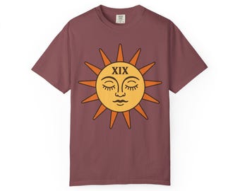 May include: A maroon t-shirt featuring a stylized sun graphic. The sun has a yellow face with closed eyes and the Roman numeral XIX. The sun's rays are orange. The shirt is a short-sleeved crew neck.