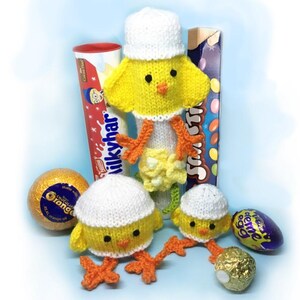 Peut inclure: Plusieurs poussins jaunes tricotés avec des chapeaux blancs et des pieds oranges, accompagnés de diverses friandises au chocolat. Les poussins sont disposés avec une barre Milky Way, une boîte de Smarties et d'autres œufs et bonbons au chocolat, sur fond bleu clair.