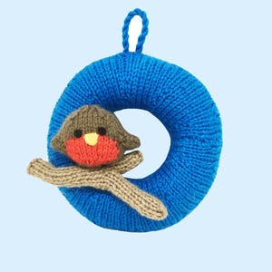 May include: A knitted Christmas ornament featuring a blue wreath with a loop for hanging. A small, knitted bird with a red breast, brown head, and tan wings is attached to the wreath. The background is a light blue.