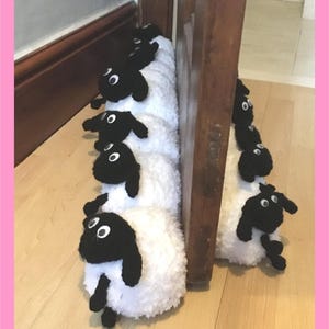 May include: A novelty doorstop featuring a row of sheep. Each sheep has a white body, black face, ears, and legs. The doorstop is positioned against a wooden door. The image has a light pink border.