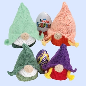 Peut inclure: Quatre gnomes tricotés en vert, pêche et violet avec des accents blancs, beiges et rouges. Deux œufs en chocolat sont placés entre les gnomes. Les gnomes ont des chapeaux pointus et des nez ronds. Le fond est bleu clair.