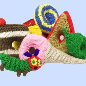May include: A selection of knitted food items and a caterpillar, including a pink doughnut, ice cream cone, cake, and a green caterpillar. The items are arranged on a light blue background, showcasing a playful, handmade aesthetic.