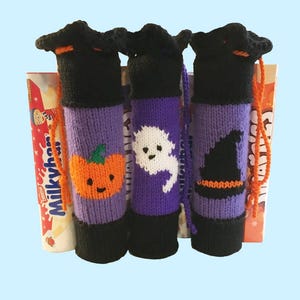 Könnte beinhalten: Drei Halloween-Flaschenhüllen aus Strick. Jede Hülle ist schwarz, lila und orange und zeigt einen Kürbis, einen Geist und einen Hexenhut. Die Hüllen sind zylindrisch und haben oben einen Kordelzugverschluss.