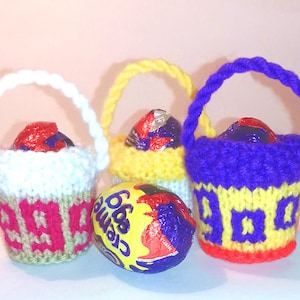 May include: Three knitted Easter baskets in white, yellow, and purple, each with a handle and filled with a chocolate egg. A fourth egg, wrapped in yellow foil, sits in front. The baskets have decorative patterns.