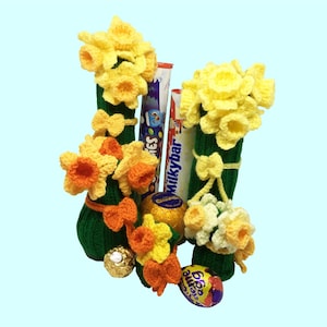 May include: A handcrafted Easter arrangement featuring knitted green vases adorned with yellow and orange crochet daffodils and bows. The arrangement includes chocolate bars, a gold-wrapped chocolate, and a decorated Easter egg.