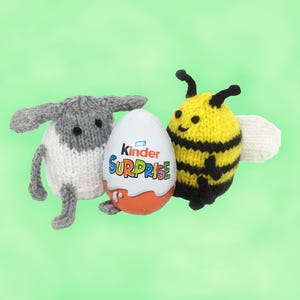 May include: A knitted sheep, a Kinder Surprise egg, and a knitted bee are displayed against a green backdrop. The sheep is grey and white, the egg is white with orange at the base, and the bee is yellow and black. The egg has the words "Kinder Surprise" on it.