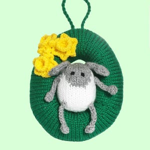 May include: A handmade Easter decoration featuring a green knitted oval with a loop for hanging. A knitted bunny with gray and white details is centered on the oval, with yellow knitted flowers on the left side. The background is a light green.