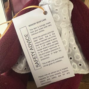 Pode incluir: Um urso de pelúcia bordô vestindo um vestido branco com detalhes de ilhós. Uma etiqueta com o texto "SAFETY ADVICE" e "MEMORY BEAR CARE" está anexada. A etiqueta fornece informações de segurança e instruções de cuidados.