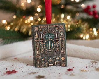 May include: A decorative Christmas tree ornament shaped like a book, with the title "A Christmas Carol" on the cover. The ornament is dark green with gold detailing and a red ribbon. The background features Christmas lights and a soft, white surface.