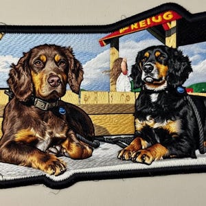 Könnte beinhalten: Gestickter Aufnäher mit zwei Hunden, einem braunen und einem schwarzen, die liegen. Die Hunde tragen Halsbänder und Leinen. Der Hintergrund zeigt eine Holzkonstruktion und das Wort "REIUG". Der Aufnäher hat einen schwarzen Rand.