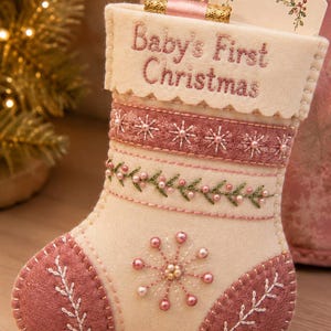 May include: A cream-coloured felt Christmas stocking featuring the embroidered text Baby's First Christmas. The stocking is decorated with pink felt accents, intricate floral embroidery, and small pink beads arranged in a starburst pattern.