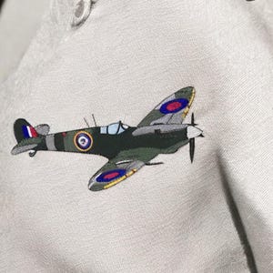 Può includere: Immagine ricamata di un aereo verde scuro e grigio con dettagli rossi, blu e gialli. L'aereo ha un'elica ed è posto su uno sfondo grigio chiaro. Il disegno è dettagliato e sembra essere cucito.