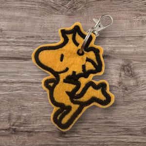 Puede incluir: Un llavero de fieltro amarillo con un personaje de dibujos animados con contornos y detalles negros. El llavero tiene un cierre plateado para sujetarlo a llaves o bolsos. El personaje está en una pose juguetona.