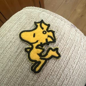 May include: A yellow felt patch of Woodstock, the Peanuts character, with black edging. The patch is displayed on a textured, off-white surface. A fun, cartoon-themed item.