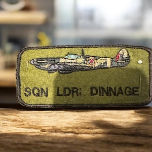 May include: A rectangular embroidered patch with a detailed image of a military aircraft in green, black, and tan. The patch includes the text "SQN LDR: DINNAGE" in black lettering on a green background.