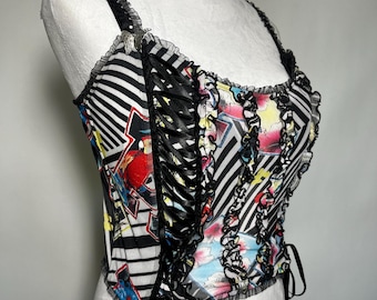 May include: A patterned corset top with black and white stripes, floral and geometric designs, and black ribbon detailing. The top has ruffled edges and thin straps. The corset is displayed on a white mannequin.