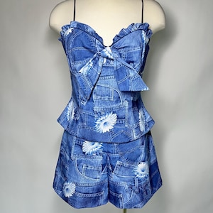 May include: A two-piece outfit with a denim print on a mannequin. The top has a ruffled neckline, spaghetti straps, and a large bow. The shorts have a high waist. White daisy floral patterns are scattered across the blue denim fabric.