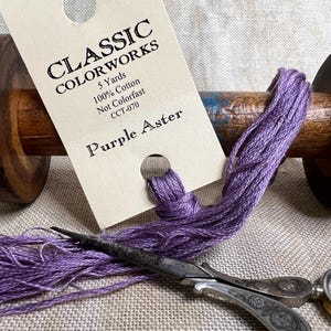 May include: A spool of purple embroidery floss labeled "Classic Colorworks Purple Aster" with a tag that reads "5 Yards 100% Cotton Not Colorfast CCT-070".