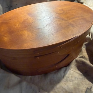 May include: An oval-shaped wooden box with a smooth, polished finish in a warm medium-brown tone. The container features a fitted lid and a side wall constructed with overlapping wood bands secured by small, evenly spaced metal fasteners.