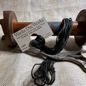 May include: A wooden thread spool with black thread and a pair of decorative scissors. A tag reads "Weeks Dye Works" and "1304 Onyx". The spool and scissors rest on a textured, neutral-colored surface.