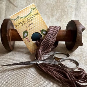 May include: A wooden spool with a brown thread wound around it. A small card with the text "Romy's Creations Colors and Threads Coffee" is next to the spool. A pair of antique scissors is on top of the thread.