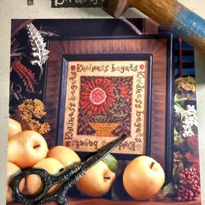 May include: A framed cross-stitch sampler with the text "Kindness begets" and a floral design. A pair of ornate scissors rests in front of the frame, with several pears and other decorative elements. The image is set against a backdrop of a wooden hammer and a page with the text "Birds of".