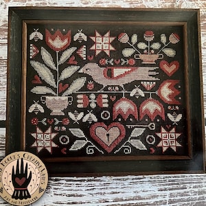 May include: A cross-stitch pattern featuring a red bird, hearts, flowers, and stars on a black background. The pattern is framed in a dark wood frame.