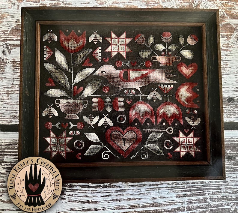 May include: A cross-stitch pattern featuring a red bird, hearts, flowers, and stars on a black background. The pattern is framed in a dark wood frame.