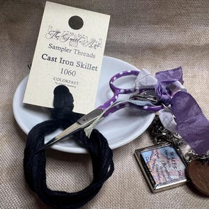May include: A white ceramic dish with a black embroidery floss wound around it. A pair of purple and white polka dot scissors with a silver handle is resting on the dish. A small tag with the text "The Gentle Art Sampler Threads Cast Iron Skillet 1060 COLORFAST" is visible.