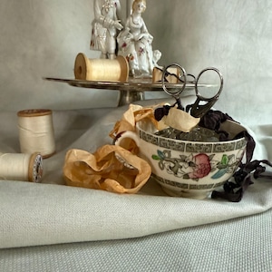 May include: A ceramic teacup with a floral pattern and geometric border containing metal scissors and ribbons, accompanied by wooden spools of thread and a silver pedestal stand featuring two porcelain figurines of a couple in period attire.