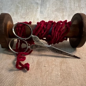 May include: A wooden spool with a red fuzzy yarn wrapped around it. A pair of silver scissors with ornate handles are lying on top of the yarn.