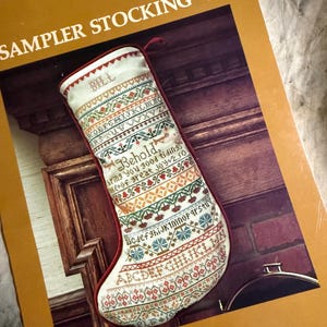 May include: A cream-colored, embroidered Christmas stocking with a red trim, resting on a book titled "Sampler Stocking V." The stocking features colorful cross-stitch patterns, including letters and floral designs. The word "Bill" is stitched at the top.