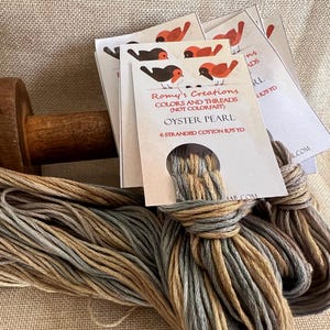 May include: A skein of six-strand cotton embroidery floss in a color called "Oyster Pearl". The floss is a blend of light brown, green, and gray. The skein is wrapped around a wooden spool. The floss is labeled "Romy's Creations Colors and Threads (Not Colorfast) Oyster Pearl 6 Stranded Cotton 875 yd."