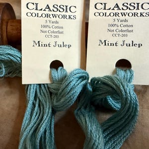 May include: Two skeins of mint green embroidery floss with tags that read "CLASSIC COLORWORKS" and "Mint Julep." Each skein is 5 yards of 100% cotton. A wooden spool is visible in the background.