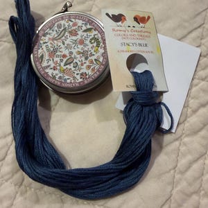 May include: A round metal tin with a floral pattern and the text Maison Sajou Paris, alongside a skein of dark blue embroidery thread attached to a paper tag labeled Romy's Creations, Stacy's Blue, 6 stranded cotton, and not colorfast.