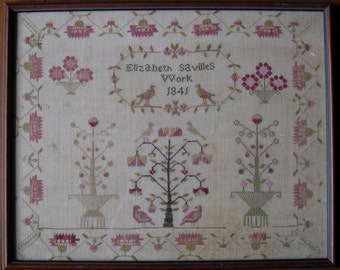 Op de afbeelding: Een ingelijst borduurwerk met een witte achtergrond en roze en groene borduursels. Het borduurwerk toont drie bomen, elk met een ander ontwerp. De tekst "Elizabeth Savilles Work 1841" is geborduurd in het midden van het borduurwerk.