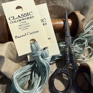 May include: Close-up of craft supplies including a skein of light blue thread, vintage scissors, and a wooden spool. The thread is attached to a tag that reads "CLASSIC COLORWORKS Barrel Cactus". The scissors have ornate handles.