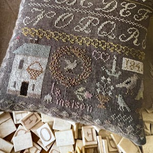 May include: A decorative, embroidered pillow with a dark gray background. The pillow features a house, a wreath, and the text "Le maison". The design includes stitched letters and floral accents. A collection of letter tiles and wooden rings are in the foreground.