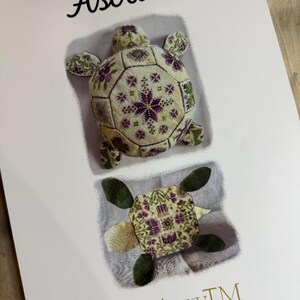 May include: A print featuring two turtle designs with floral patterns in purple, green, and cream. The word "Astrid" is written in cursive at the top. The bottom right corner has the text "AuryTM".