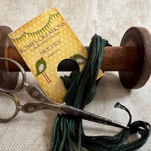 May include: A wooden thread spool with dark green thread, a pair of silver scissors, and a card labeled "Mistletoe" with bird illustrations. The card is yellow with white polka dots.