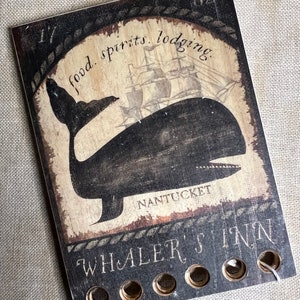 May include: A rectangular wooden sign featuring a black silhouette of a whale, a sailing ship, and the text "food. spirits. lodging." and "WHALER'S INN NANTUCKET". The sign has a distressed finish and five circular holes along the bottom edge.