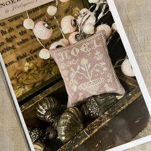 May include: A cross stitch pattern for a pillow with the words "Noel Tuffet" in a pink and white color scheme. The pillow is surrounded by decorative ornaments in a wooden box.